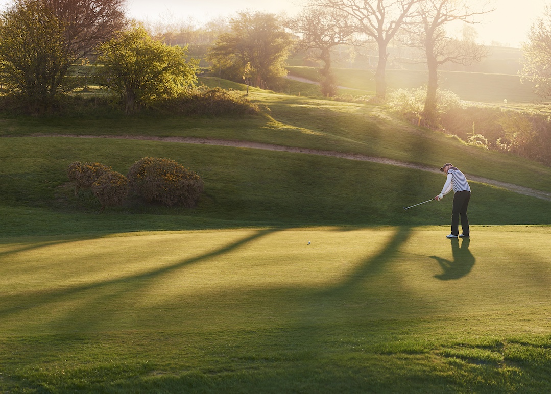 a lone golfer putts on the green , the low sun is coming from behind him and flaring to camera.